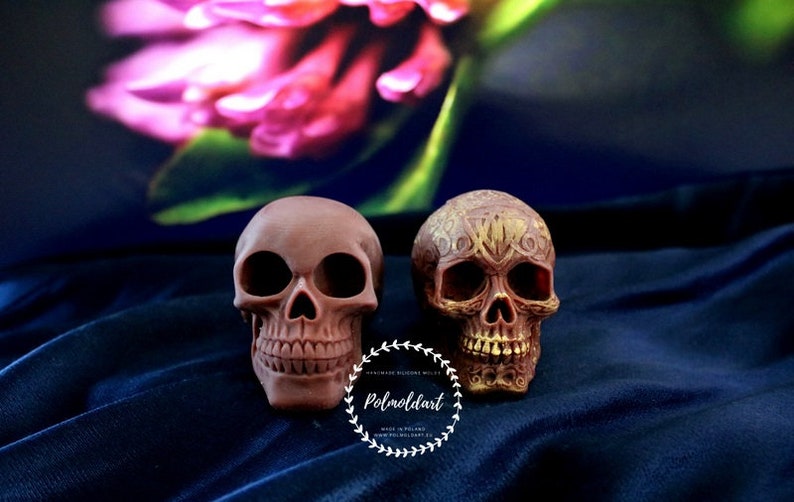 May include: Two brown skull figurines. One is plain, the other is decorated with an ornate gold design. The skulls are set against a dark blue background with a blurred floral element. The text "Polmoldart" is visible.