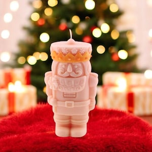 May include: A pink candle in the shape of a nutcracker soldier, adorned with a gold crown and belt, rests on a red surface. The background shows a Christmas tree with bokeh lights and wrapped gifts, creating a festive holiday scene.