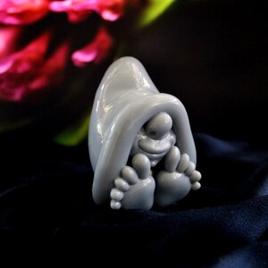 May include: A whimsical gray figurine of a creature with a hooded cloak, smiling face, and large feet. The sculpture is set against a dark blue fabric with a blurred floral background.