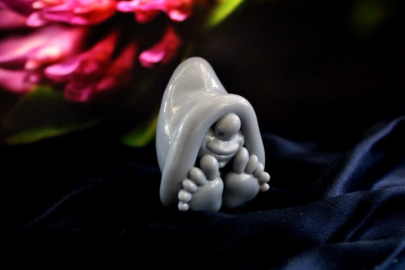 May include: A whimsical gray figurine of a creature with a hooded cloak, smiling face, and large feet. The sculpture is set against a dark blue fabric with a blurred floral background.