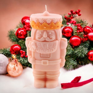 May include: A light pink candle shaped like a Nutcracker soldier, with a gold crown and intricate details. The candle is set against a backdrop of Christmas decorations, including red ornaments and evergreen branches, creating a festive holiday scene.