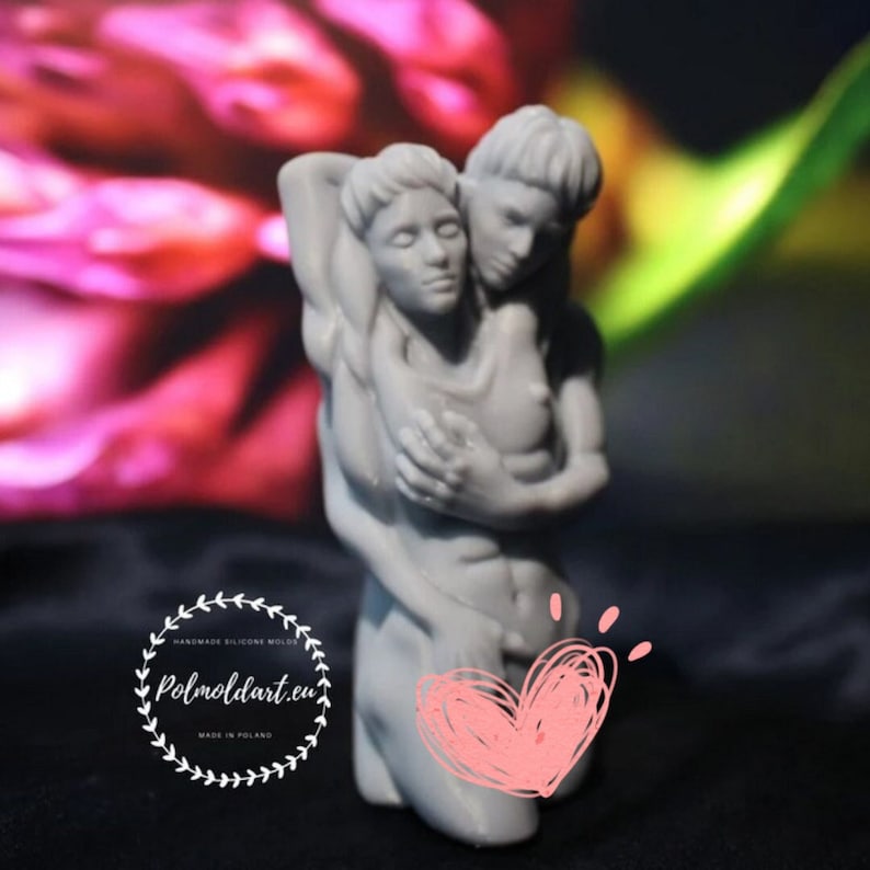 K&ouml;nnte beinhalten: Eine graue Skulptur, die zwei sich umarmende Figuren darstellt. Die Figuren sind in einem klassischen Stil mit glatten Linien und einem ruhigen Ausdruck gemei&szlig;elt. Die Skulptur steht vor einem verschwommenen rosa und roten Hintergrund.