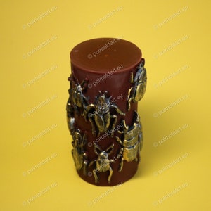 Pu&ograve; includere: Una candela cilindrica rosso scuro decorata con abbellimenti a forma di scarabeo color bronzo. La candela ha una parte superiore liscia e solida ed &egrave; posta su uno sfondo giallo. Il design presenta figure di scarabei di varie dimensioni e stili.
