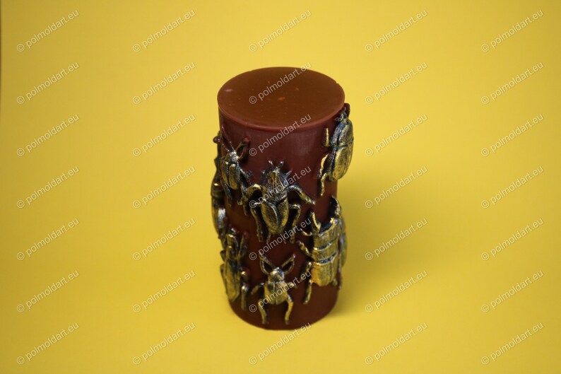 Pu&ograve; includere: Una candela cilindrica rosso scuro decorata con abbellimenti a forma di scarabeo color bronzo. La candela ha una parte superiore liscia e solida ed &egrave; posta su uno sfondo giallo. Il design presenta figure di scarabei di varie dimensioni e stili.