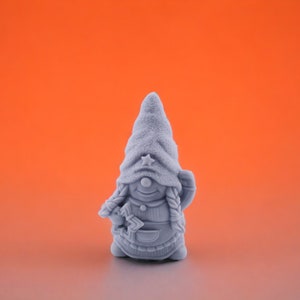 Puede incluir: Figura de gnomo gris con un sombrero alto y texturizado adornado con una estrella. El gnomo tiene trenzas, una cara sonriente y sostiene un objeto pequeño. Lleva un chaleco y un delantal, sobre un fondo naranja.