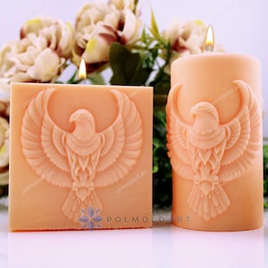 Peut inclure: Deux bougies piliers pêche, l'une carrée et l'autre cylindrique, avec un motif d'aigle sculpté. Les deux bougies sont allumées. Le fond comprend des fleurs et des sphères décoratives.