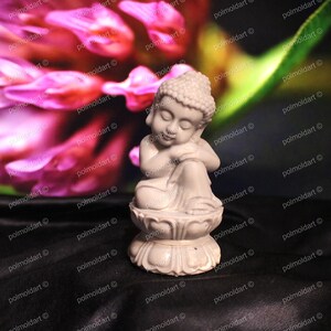 May include: A small, off-white Buddha statue in a seated position, resting on a lotus flower base. The statue has its eyes closed and is leaning on its arm. The background features a blurred pink flower and dark fabric.