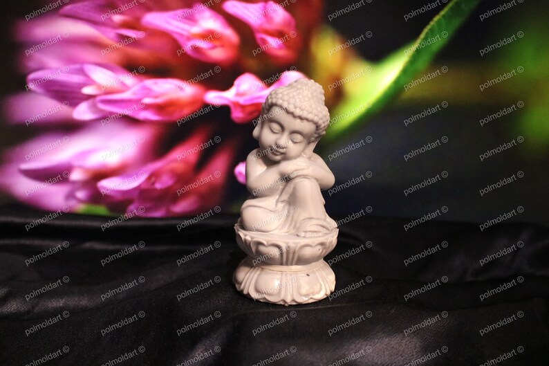 May include: A small, off-white Buddha statue in a seated position, resting on a lotus flower base. The statue has its eyes closed and is leaning on its arm. The background features a blurred pink flower and dark fabric.