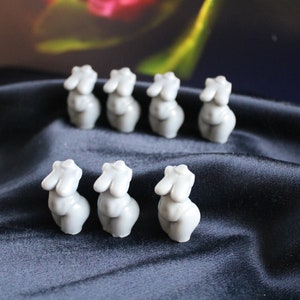 Puede incluir: Ocho peque&ntilde;as figuras de torso femenino grises est&aacute;n dispuestas sobre una tela azul oscuro y sedosa. Las figuras est&aacute;n estilizadas con formas redondeadas y superficies lisas, lo que sugiere una est&eacute;tica minimalista.