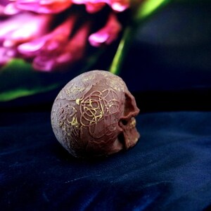 May include: A detailed, reddish-brown skull with gold-coloured, intricate designs on the top and side. The skull is positioned on a dark blue, textured fabric, with a blurred floral background.