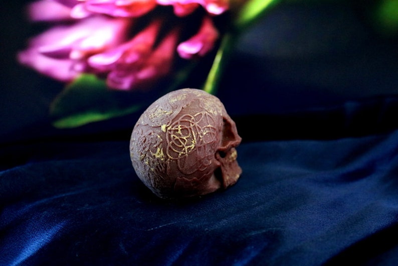 May include: A detailed, reddish-brown skull with gold-coloured, intricate designs on the top and side. The skull is positioned on a dark blue, textured fabric, with a blurred floral background.