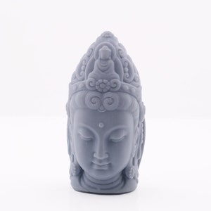 May include: A gray statue of a serene Buddha head with closed eyes, intricate hair details, and a detailed crown. The sculpture is a light gray color and stands on a white background.