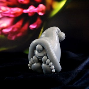 May include: A gray figurine of a whimsical creature wearing a hat with a pom-pom, with its feet visible. The figure is set against a dark background with a blurred floral design. The figurine is made of a smooth material.