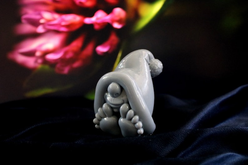 May include: A gray figurine of a whimsical creature wearing a hat with a pom-pom, with its feet visible. The figure is set against a dark background with a blurred floral design. The figurine is made of a smooth material.