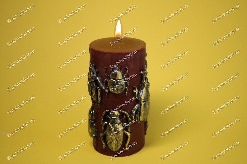 Pu&ograve; includere: Una candela a colonna bordeaux con decorazioni a scarabeo color bronzo. La candela &egrave; accesa, con una fiamma in cima. Lo sfondo &egrave; giallo pieno.