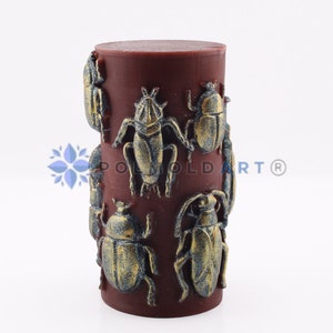 Pu&ograve; includere: Una candela cilindrica bordeaux con disegni di scarabei dorati e blu in rilievo. La candela presenta un motivo ripetuto di figure di insetti dettagliate, creando un elemento decorativo unico. Un elemento decorativo per la casa.