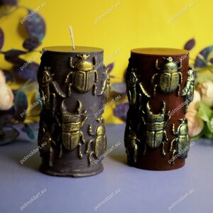 Pu&ograve; includere: Due candele a colonna decorative. Una &egrave; viola scuro, l'altra marrone rossiccio. Entrambe sono decorate con abbellimenti a forma di scarabeo dorato. Le candele hanno un aspetto rustico e strutturato e sono su uno sfondo floreale giallo e viola.