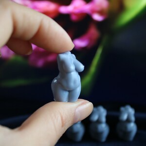 Puede incluir: Una peque&ntilde;a figura azul claro de una forma femenina se sostiene entre los dedos. La figura tiene rasgos redondeados y es una escultura en miniatura. Varias figuras similares son visibles en el fondo, con un fondo floral borroso.