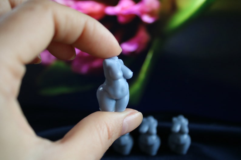 Puede incluir: Una peque&ntilde;a figura azul claro de una forma femenina se sostiene entre los dedos. La figura tiene rasgos redondeados y es una escultura en miniatura. Varias figuras similares son visibles en el fondo, con un fondo floral borroso.