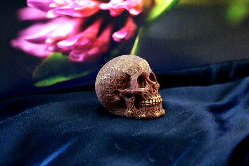 May include: A detailed, brown skull figurine with intricate gold patterns. The skull is resting on a dark blue surface, with a blurred floral background. The skull's design suggests a gothic or fantasy theme.