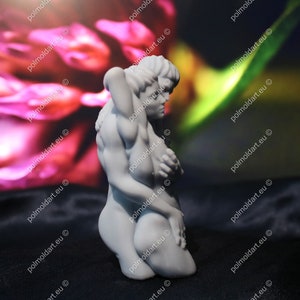 K&ouml;nnte beinhalten: Eine wei&szlig;e Skulptur, die zwei sich umarmende Figuren darstellt. Eine Figur kniet, die andere umschlingt sie. Der Hintergrund ist verschwommen, rosa und gr&uuml;n, und die Skulptur steht auf einer schwarzen Oberfl&auml;che.