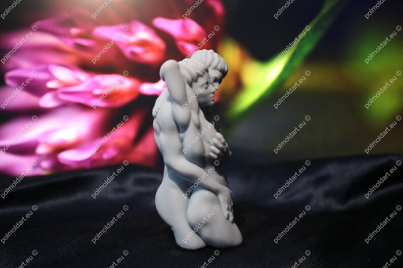 K&ouml;nnte beinhalten: Eine wei&szlig;e Skulptur, die zwei sich umarmende Figuren darstellt. Eine Figur kniet, die andere umschlingt sie. Der Hintergrund ist verschwommen, rosa und gr&uuml;n, und die Skulptur steht auf einer schwarzen Oberfl&auml;che.