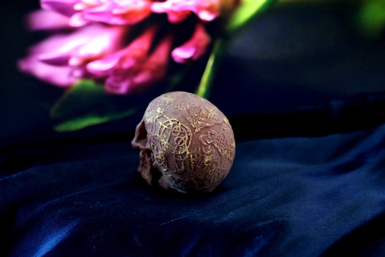 May include: A small, brown skull figurine with gold-coloured Celtic knot designs. The skull is resting on a dark blue, textured surface, with a blurred pink flower in the background. The skull is a decorative item.