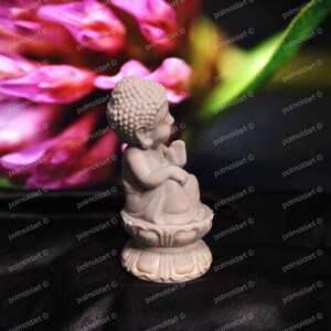 May include: A small, light gray Buddha statue seated on a lotus flower base. The statue has a serene expression and is set against a blurred background of pink flowers and green leaves. The statue is a decorative item.
