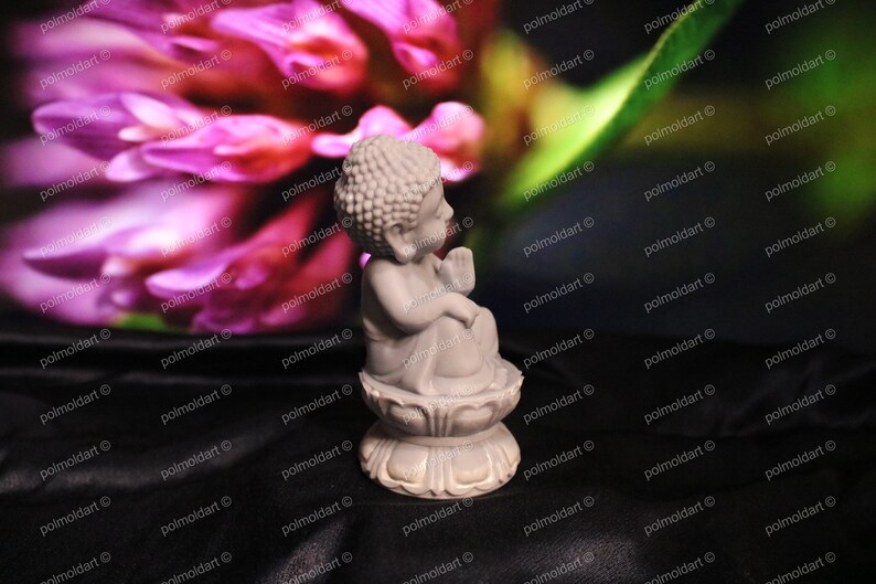 May include: A small, light gray Buddha statue seated on a lotus flower base. The statue has a serene expression and is set against a blurred background of pink flowers and green leaves. The statue is a decorative item.