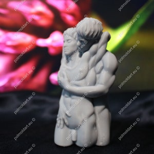 K&ouml;nnte beinhalten: Eine graue Skulptur, die zwei sich umarmende Figuren darstellt. Die Arme der m&auml;nnlichen Figur umschlie&szlig;en die weibliche Figur, ihre K&ouml;rper ber&uuml;hren sich. Die Skulptur steht vor einem verschwommenen Hintergrund aus Rosa und Gr&uuml;n.