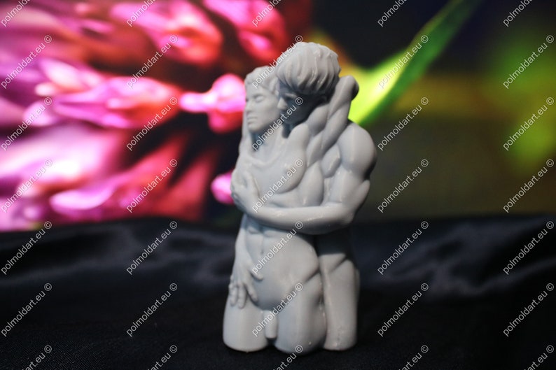 K&ouml;nnte beinhalten: Eine graue Skulptur, die zwei sich umarmende Figuren darstellt. Die Arme der m&auml;nnlichen Figur umschlie&szlig;en die weibliche Figur, ihre K&ouml;rper ber&uuml;hren sich. Die Skulptur steht vor einem verschwommenen Hintergrund aus Rosa und Gr&uuml;n.