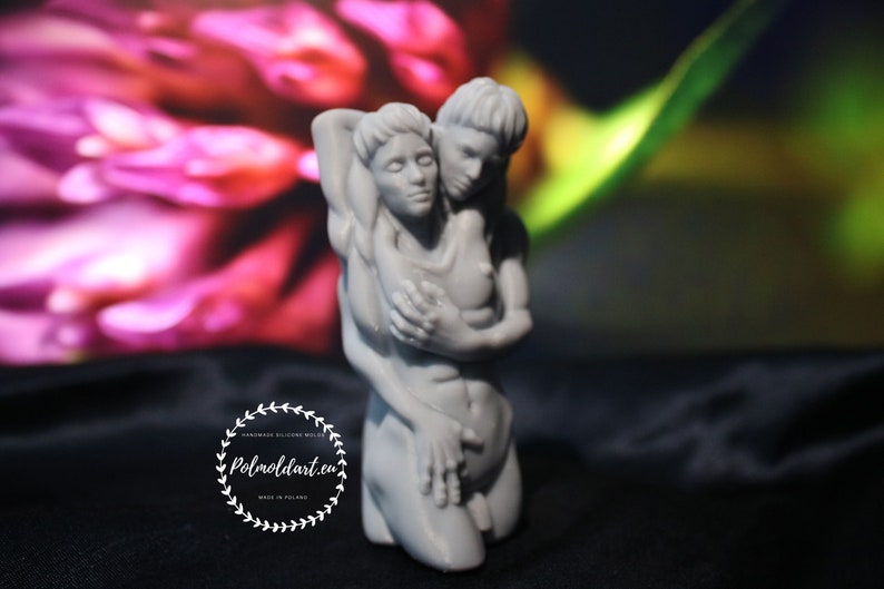K&ouml;nnte beinhalten: Eine graue Skulptur, die zwei sich umarmende Figuren darstellt. Die Figuren sind nackt und in enger Umarmung. Die Skulptur befindet sich vor einem verschwommenen Hintergrund aus rosa und gr&uuml;nen Farbt&ouml;nen. Die Skulptur tr&auml;gt den Text "Polmoldart.eu" und "Handmade silicone molds" in einem kreisf&ouml;rmigen Design.