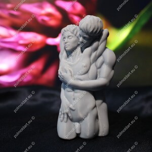 K&ouml;nnte beinhalten: Eine graue Skulptur eines sich umarmenden Paares. Die m&auml;nnliche Figur befindet sich hinter der weiblichen Figur, mit den Armen um sie gelegt. Die Skulptur steht vor einem verschwommenen Hintergrund aus Rosa und Gr&uuml;n.