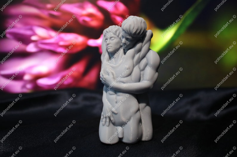 K&ouml;nnte beinhalten: Eine graue Skulptur eines sich umarmenden Paares. Die m&auml;nnliche Figur befindet sich hinter der weiblichen Figur, mit den Armen um sie gelegt. Die Skulptur steht vor einem verschwommenen Hintergrund aus Rosa und Gr&uuml;n.