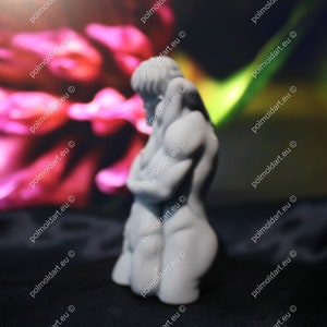 K&ouml;nnte beinhalten: Kleine, graue Skulptur einer menschlichen Figur in sitzender Position, Arme verschr&auml;nkt. Die Figur befindet sich vor einem verschwommenen Hintergrund in Rosa- und Gr&uuml;nt&ouml;nen. Die Skulptur steht auf einer schwarzen Oberfl&auml;che.