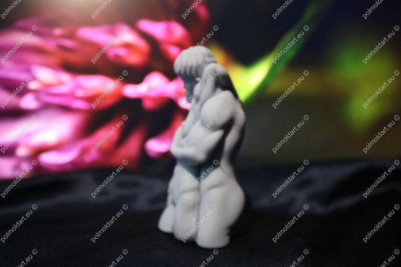 K&ouml;nnte beinhalten: Kleine, graue Skulptur einer menschlichen Figur in sitzender Position, Arme verschr&auml;nkt. Die Figur befindet sich vor einem verschwommenen Hintergrund in Rosa- und Gr&uuml;nt&ouml;nen. Die Skulptur steht auf einer schwarzen Oberfl&auml;che.