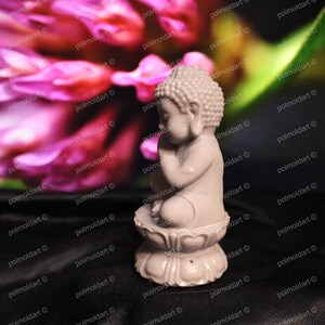 May include: A small, light gray Buddha statue in a seated position, resting on a lotus flower base. The statue is set against a blurred background of pink and green hues. The figurine is approximately 3 inches tall.