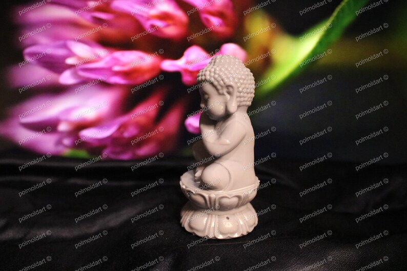 May include: A small, light gray Buddha statue in a seated position, resting on a lotus flower base. The statue is set against a blurred background of pink and green hues. The figurine is approximately 3 inches tall.