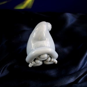 May include: A light blue ceramic gnome figurine with a pointed hat and curled feet. The gnome is resting on a dark blue, textured surface. The figurine has a glossy finish and is a whimsical decorative item.