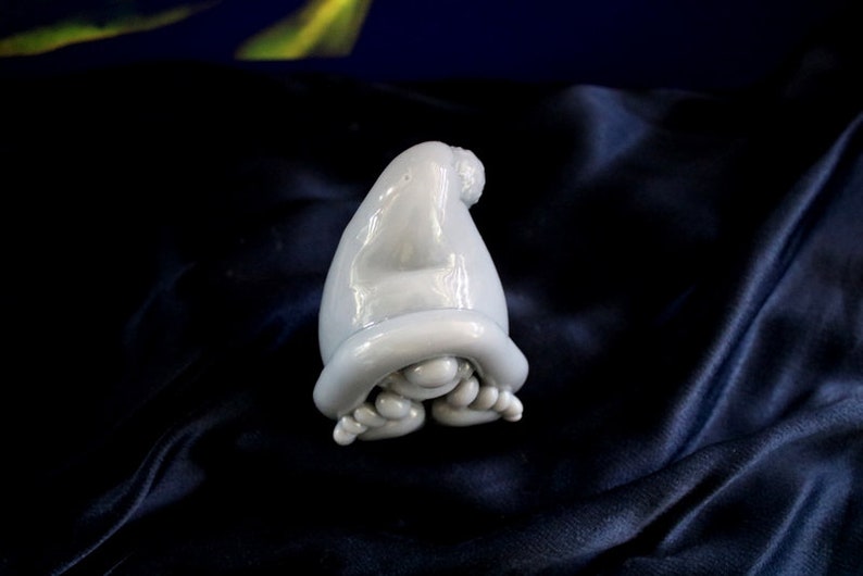 May include: A light blue ceramic gnome figurine with a pointed hat and curled feet. The gnome is resting on a dark blue, textured surface. The figurine has a glossy finish and is a whimsical decorative item.