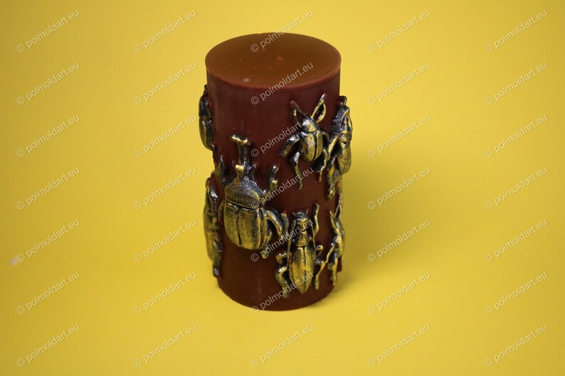 Pu&ograve; includere: Una candela cilindrica marrone scuro con abbellimenti di insetti color bronzo. La candela ha una parte superiore liscia e arrotondata ed &egrave; decorata con vari disegni dettagliati di scarabei e insetti. Lo sfondo &egrave; giallo pieno.