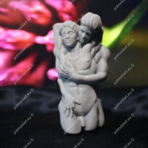 K&ouml;nnte beinhalten: Eine graue Skulptur, die zwei sich umarmende Figuren darstellt. Eine Figur wiegt die andere, mit Armen um den Oberk&ouml;rper. Die Skulptur steht vor einem verschwommenen rosa-gr&uuml;nen Hintergrund.