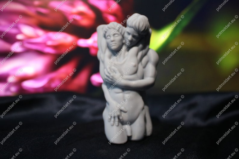 K&ouml;nnte beinhalten: Eine graue Skulptur, die zwei sich umarmende Figuren darstellt. Eine Figur wiegt die andere, mit Armen um den Oberk&ouml;rper. Die Skulptur steht vor einem verschwommenen rosa-gr&uuml;nen Hintergrund.