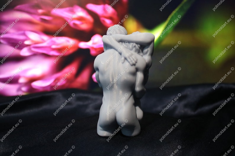 K&ouml;nnte beinhalten: Eine graue Skulptur, die zwei sich umarmende Figuren darstellt. Die Figuren sind nackt und eng umschlungen, wobei eine Figur die andere st&uuml;tzt. Der Hintergrund ist verschwommen mit rosa und gr&uuml;nen Farbt&ouml;nen.