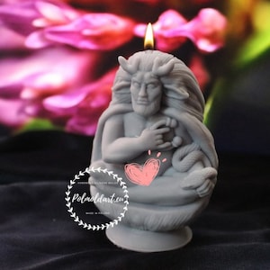 Può includere: Una candela grigia a forma di figura mitologica con corna, barba e capelli lunghi. La candela è accesa, con una fiamma in cima. Un serpente è avvolto attorno alle gambe della figura. La candela ha il testo "Polmoldart.eu" e "Made in Poland".