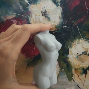 May include: A light gray, small sculpture of a female torso. The sculpture is smooth and detailed, with defined curves and anatomical features. The background is a colorful painting with white, red, and green brushstrokes.