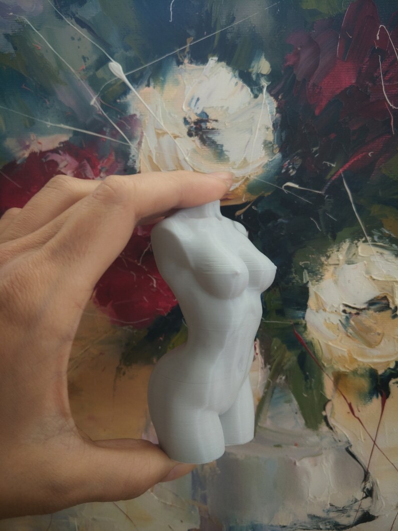 May include: A light gray, small sculpture of a female torso. The sculpture is smooth and detailed, with defined curves and anatomical features. The background is a colorful painting with white, red, and green brushstrokes.