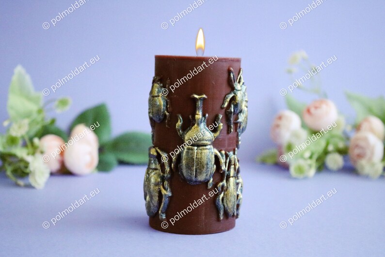 Pu&ograve; includere: Una candela a colonna marrone con una fiamma tremolante. La candela &egrave; decorata con abbellimenti a forma di scarabeo color bronzo. Lo sfondo &egrave; un viola tenue con accenti floreali, creando un oggetto decorativo unico.