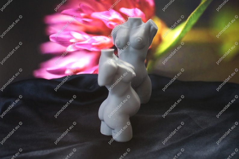 May include: Two grey, sculpted female torso statues. The statues are positioned in front of a blurred background with pink and green hues. The statues are on a black surface.