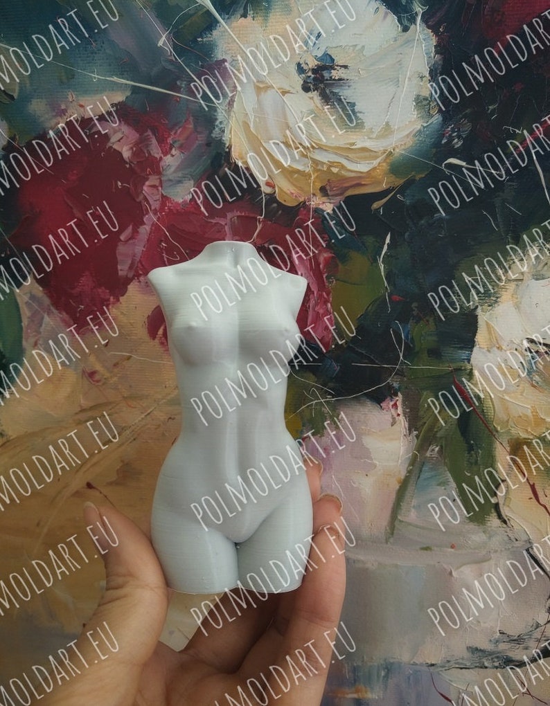 May include: A small, white, sculpted female torso figurine. The figure is held in a hand against a backdrop of a colorful, abstract painting. The figurine is a detailed representation of the female form.